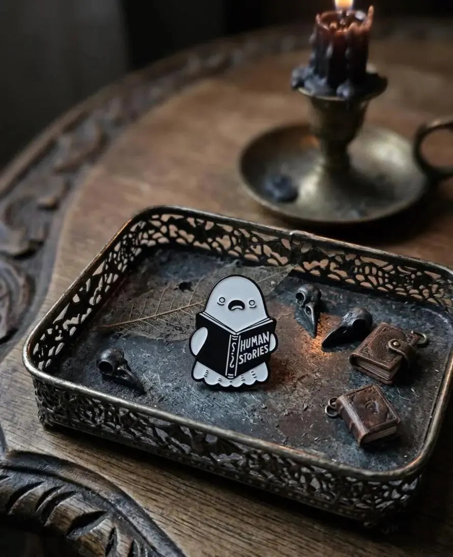 A hand holds a small enamel pin shaped like a sleeping cartoon character reading a book labeled 'HUMAN STORIES' against a softly blurred indoor tabletop scene with flowers, glasses, and a small clock. Human Stories Ghost Enamel Pin - Cute Funny Reading Ghost Pin, Bookworm Gifts - Spooky Introvert Gift, Spooky Season Gifts, Dark Academia