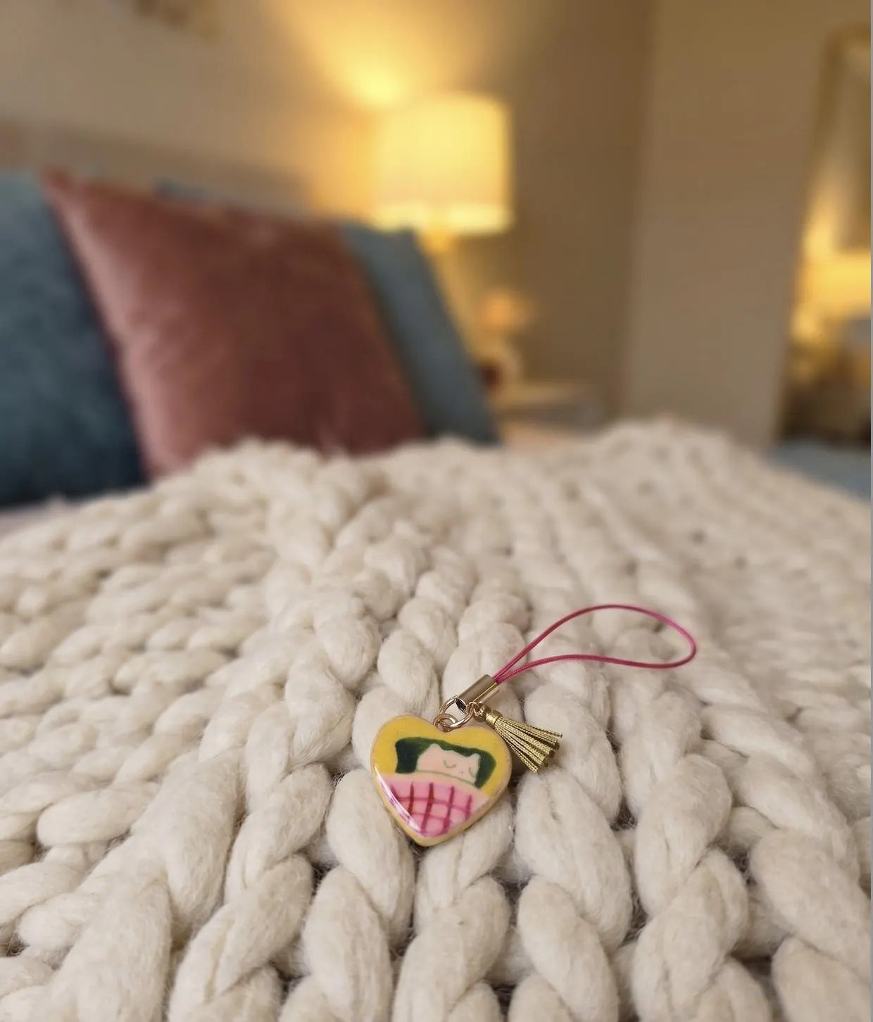 A hand with pink manicured nails holds a small heart-shaped charm with a pink strap. The charm features a yellow face with black hair and a pink-and-white striped skirt, set against a softly blurred bedroom background. Professional Cozy Napper Cat Phone Charm, Nap Lovers Gift, Vintage pendants and Charms, Jewellery Accessory, Introvert Keychain, Lazy Vibes
