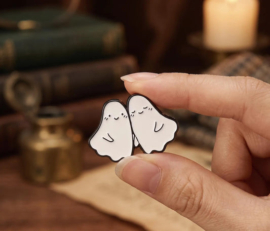 A hand holds a pair of small white ghost-shaped enamel pins with simple black line drawings, set against a cozy, blurred background of books, a cup of tea, and a feather quill. Forever Ghost Besties Enamel Pin | Soft Goth Aesthetic Halloween Pin | RIP Ghosts Spooky Lovers Gift | Cute Matching Best Friend Lapel Pin best friend pin, matching bff pins, cute ghost pin, halloween pin, soft goth pin, friendship gift, bestie gift, spooky cute gift, backpack pin, matching pins, gift for bff, hard enamel pin, kawaii