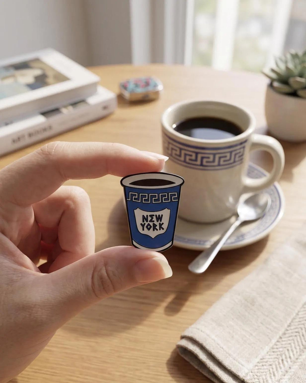 A hand holds a small enamel pin shaped like a blue coffee cup with the words 'NEW YORK' on it, set against a warm, softly blurred cafe table scene with a cup of coffee, spoon, and books. Iconic NYC Blue Coffee Cup Enamel Pin - Anthora Greek Cup Pin, New York City Souvenir, Retro Bodega Aesthetic, Coffee Lover Gifts, Java NYC Coffee Cup Enamel Pin, Iconic Anthora Greek Cup Badge, New York Souvenir Gift, Retro Bodega Aesthetic Pin, Coffee Lover Backpack Flair