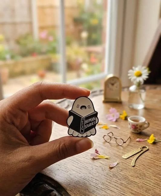 A hand holds a small enamel pin shaped like a sleeping cartoon character reading a book labeled 'HUMAN STORIES' against a softly blurred indoor tabletop scene with flowers, glasses, and a small clock. Human Stories Ghost Enamel Pin - Cute Funny Reading Ghost Pin, Bookworm Gifts - Spooky Introvert Gift, Spooky Season Gifts, Dark Academia