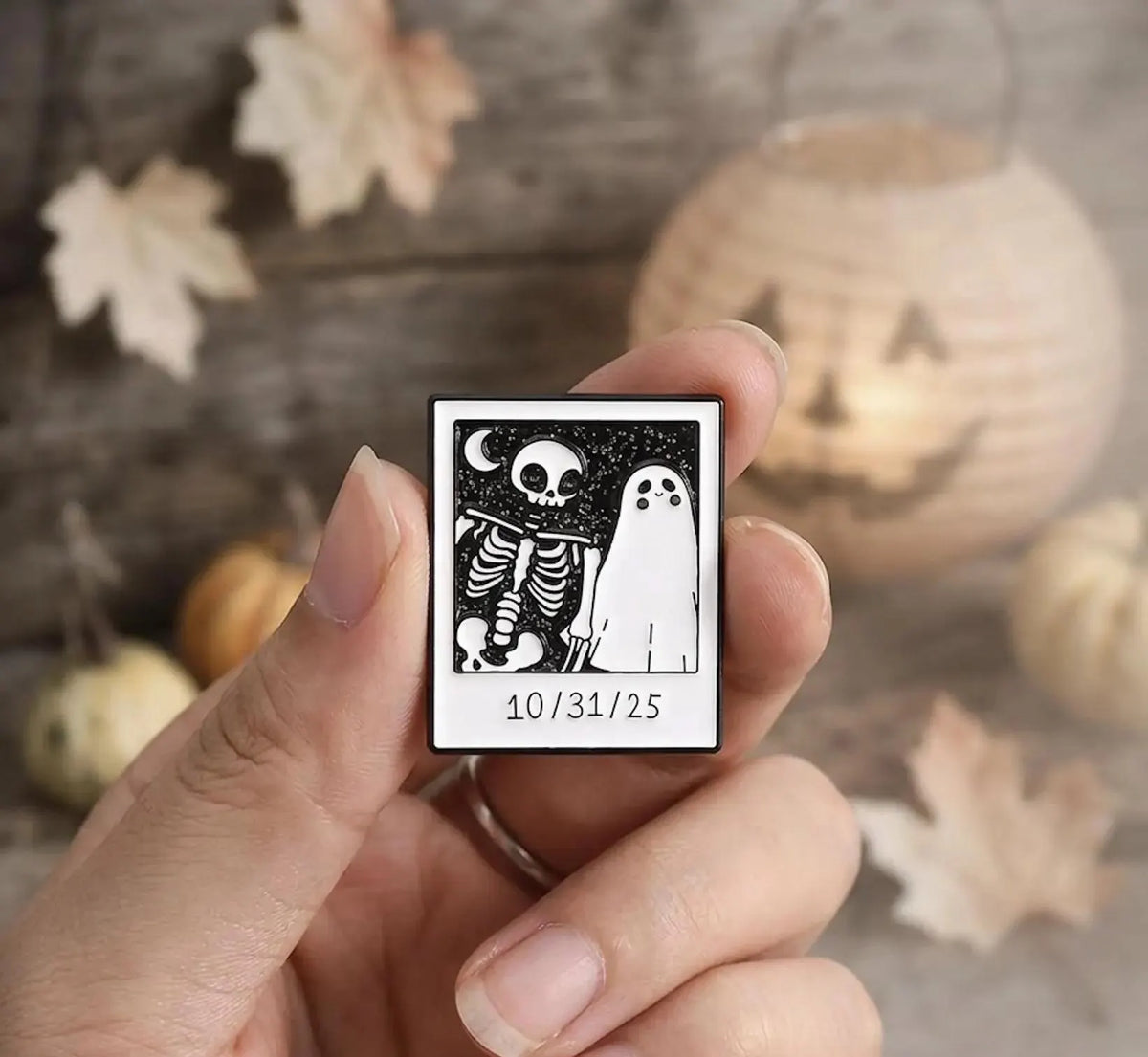 A canvas tote bag laid on a wooden surface with dried leaves and a knit blanket nearby. On the bag are several small enamel pins: a square black-and-white skeleton-and-ghost design with the date '10. Spooky Ghost and Skeleton Enamel Pin - Cute Halloween Polaroid Lapel Pin - Aesthetic Gothic Jewelry - Retro Spooky Season Gift for friends