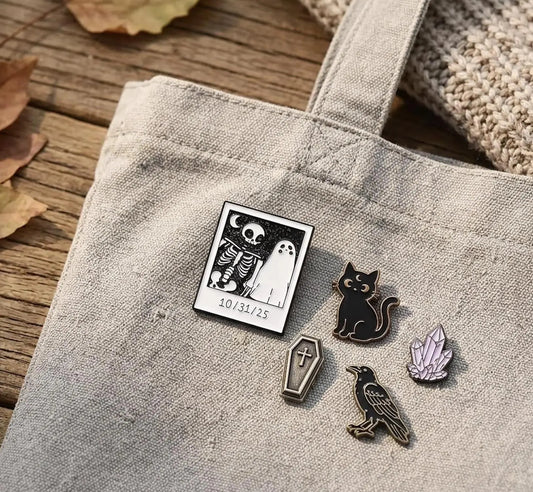 A canvas tote bag laid on a wooden surface with dried leaves and a knit blanket nearby. On the bag are several small enamel pins: a square black-and-white skeleton-and-ghost design with the date '10. Spooky Ghost and Skeleton Enamel Pin - Cute Halloween Polaroid Lapel Pin - Aesthetic Gothic Jewelry - Retro Spooky Season Gift for friends