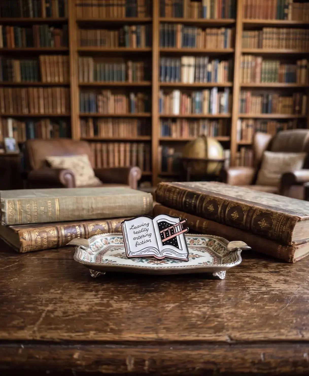 A hand holding a small enamel pin shaped like an open book with the phrase 'Zwilling really enjoying fiction' on the left page. The background shows a beige canvas tote bag and a blurred wooden surface with old books. Fiction Lover Enamel Pin - Cute Book Lover Pin, Bibliophile Gift for Readers, Bookish Accessories, Book Lover Gifts, Bookworm gifts, Fiction