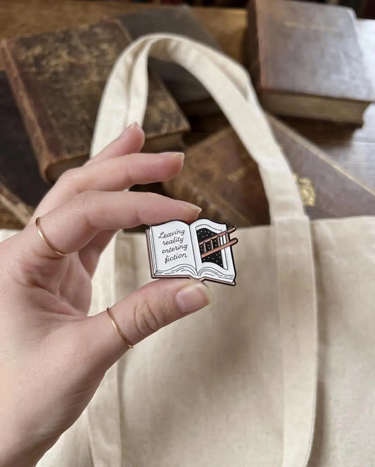 A hand holding a small enamel pin shaped like an open book with the phrase 'Zwilling really enjoying fiction' on the left page. The background shows a beige canvas tote bag and a blurred wooden surface with old books. Fiction Lover Enamel Pin - Cute Book Lover Pin, Bibliophile Gift for Readers, Bookish Accessories, Book Lover Gifts, Bookworm gifts, Fiction
