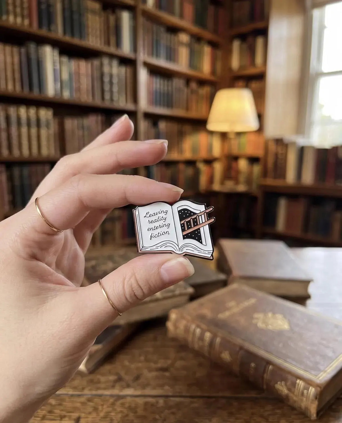 A hand holding a small enamel pin shaped like an open book with the phrase 'Zwilling really enjoying fiction' on the left page. The background shows a beige canvas tote bag and a blurred wooden surface with old books. Fiction Lover Enamel Pin - Cute Book Lover Pin, Bibliophile Gift for Readers, Bookish Accessories, Book Lover Gifts, Bookworm gifts, Fiction