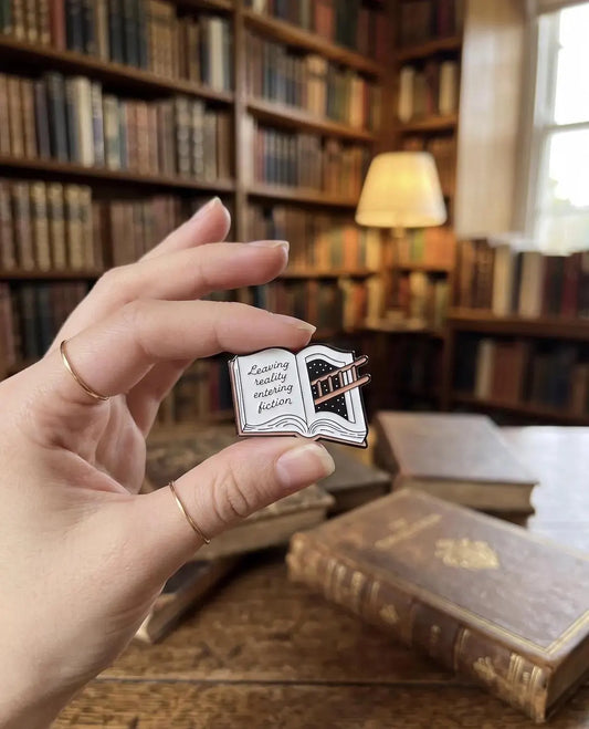 A hand holding a small enamel pin shaped like an open book with the phrase 'Zwilling really enjoying fiction' on the left page. The background shows a beige canvas tote bag and a blurred wooden surface with old books. Fiction Lover Enamel Pin - Cute Book Lover Pin, Bibliophile Gift for Readers, Bookish Accessories, Book Lover Gifts, Bookworm gifts, Fiction