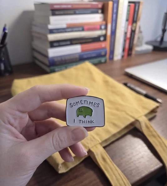 A hand holds a small enamel pin with a green cartoon creature and the text 'SOMETIMES I THINK' in front of a yellow tote bag and a blurred desk background with books. Cute Frog Enamel Pin, “Sometimes I Think” Meme Pin, Funny Relatable Gift, Mental Health Pin, Kawaii Green Pin, ADHD Pin, Gift for Friends