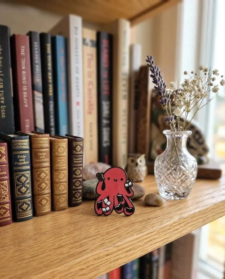 A red octopus-shaped enamel pin with black outlines and white accents, displayed on a wooden bookshelf next to books and a small glass vase with dried flowers. Red Octopus Reading Book Enamel Pin, Cute Bookworm Brooch, Nerdy Library Jewelry, Academic Animal Badge, Reader Gift, Teacher Appreciation