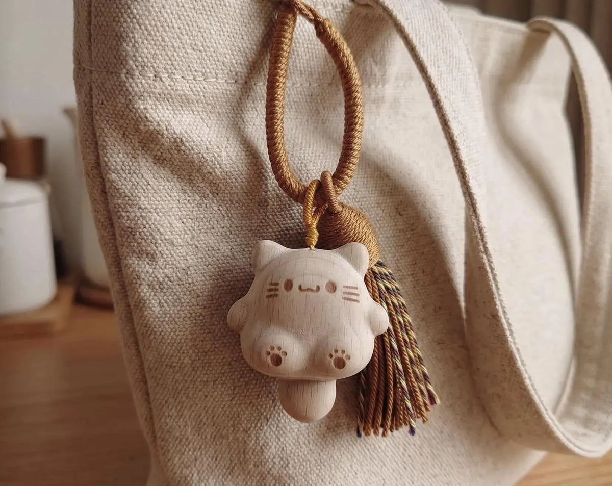 A hand holding a small carved wooden cat charm keychain with a brown braided cord and tassel against a plain light background. Handmade Wooden Cat Charm - Cute Kawaii Cat Bag Pendant, Minimalist Wood Keychain, Car Decor, Phone Strap, Unique Gift for Cat Lovers, Gifts