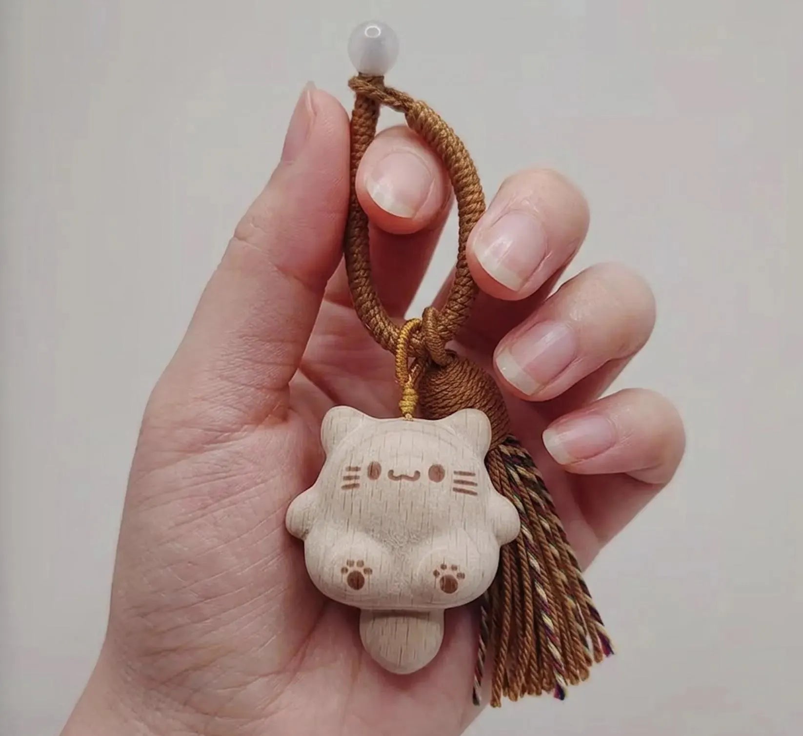 A hand holding a small carved wooden cat charm keychain with a brown braided cord and tassel against a plain light background. Handmade Wooden Cat Charm - Cute Kawaii Cat Bag Pendant, Minimalist Wood Keychain, Car Decor, Phone Strap, Unique Gift for Cat Lovers, Gifts