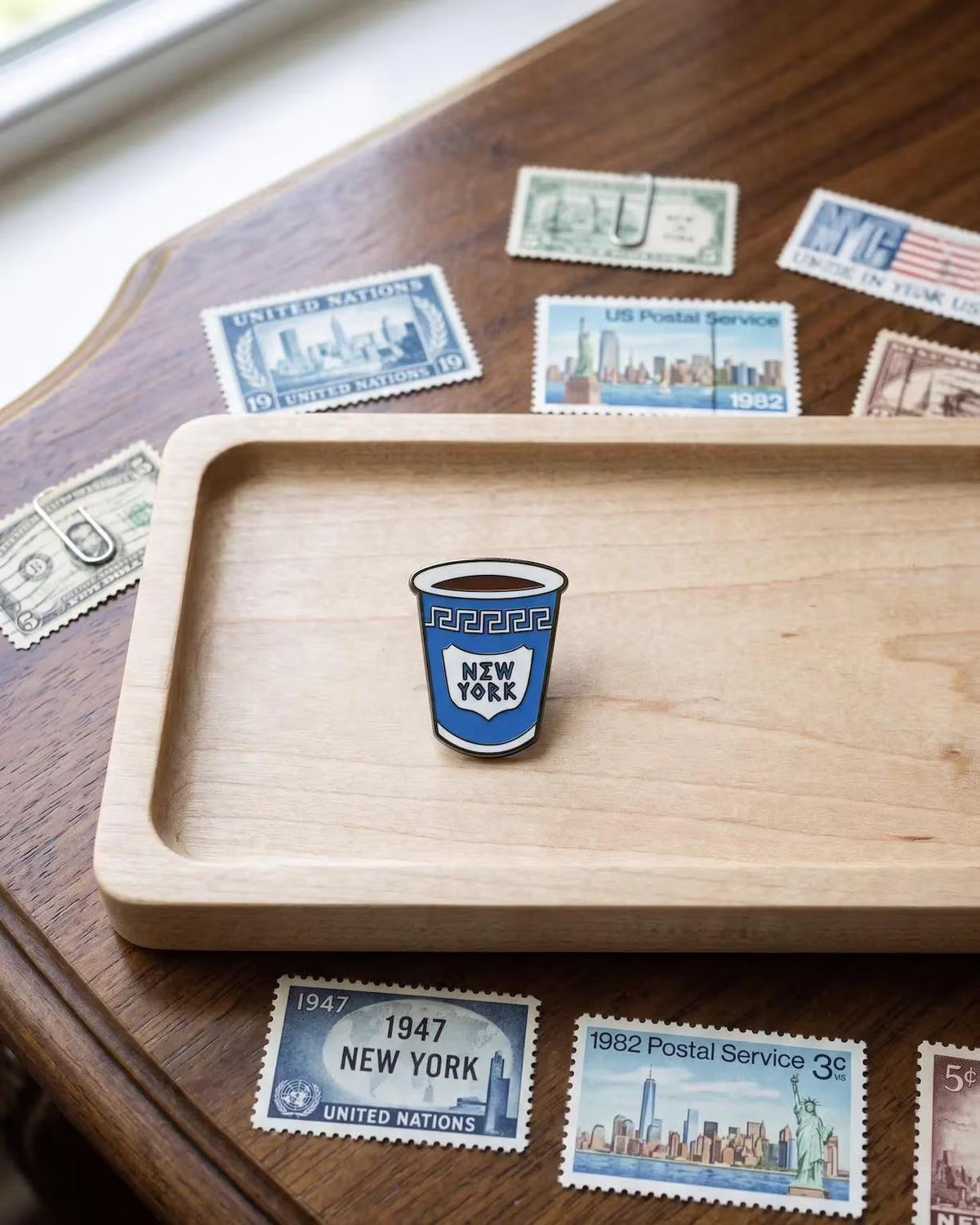 A hand holds a small enamel pin shaped like a blue coffee cup with the words 'NEW YORK' on it, set against a warm, softly blurred cafe table scene with a cup of coffee, spoon, and books. Iconic NYC Blue Coffee Cup Enamel Pin - Anthora Greek Cup Pin, New York City Souvenir, Retro Bodega Aesthetic, Coffee Lover Gifts, Java NYC Coffee Cup Enamel Pin, Iconic Anthora Greek Cup Badge, New York Souvenir Gift, Retro Bodega Aesthetic Pin, Coffee Lover Backpack Flair