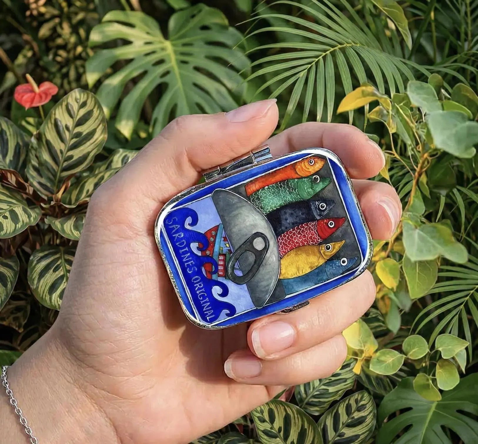 A small rectangular tin with a colorful fish illustration and the text 'PERSIMMA' sits on a light blue plate with scattered pastel candies. The background shows a softly blurred indoor setting with curtains and a table surface. Sardines Pill Case, Pocket Size Metal Mint Holder, Refillable Pill Box, Cute Kawaii Portuguese Art Accessory, Small Gifts, Pill Organizer