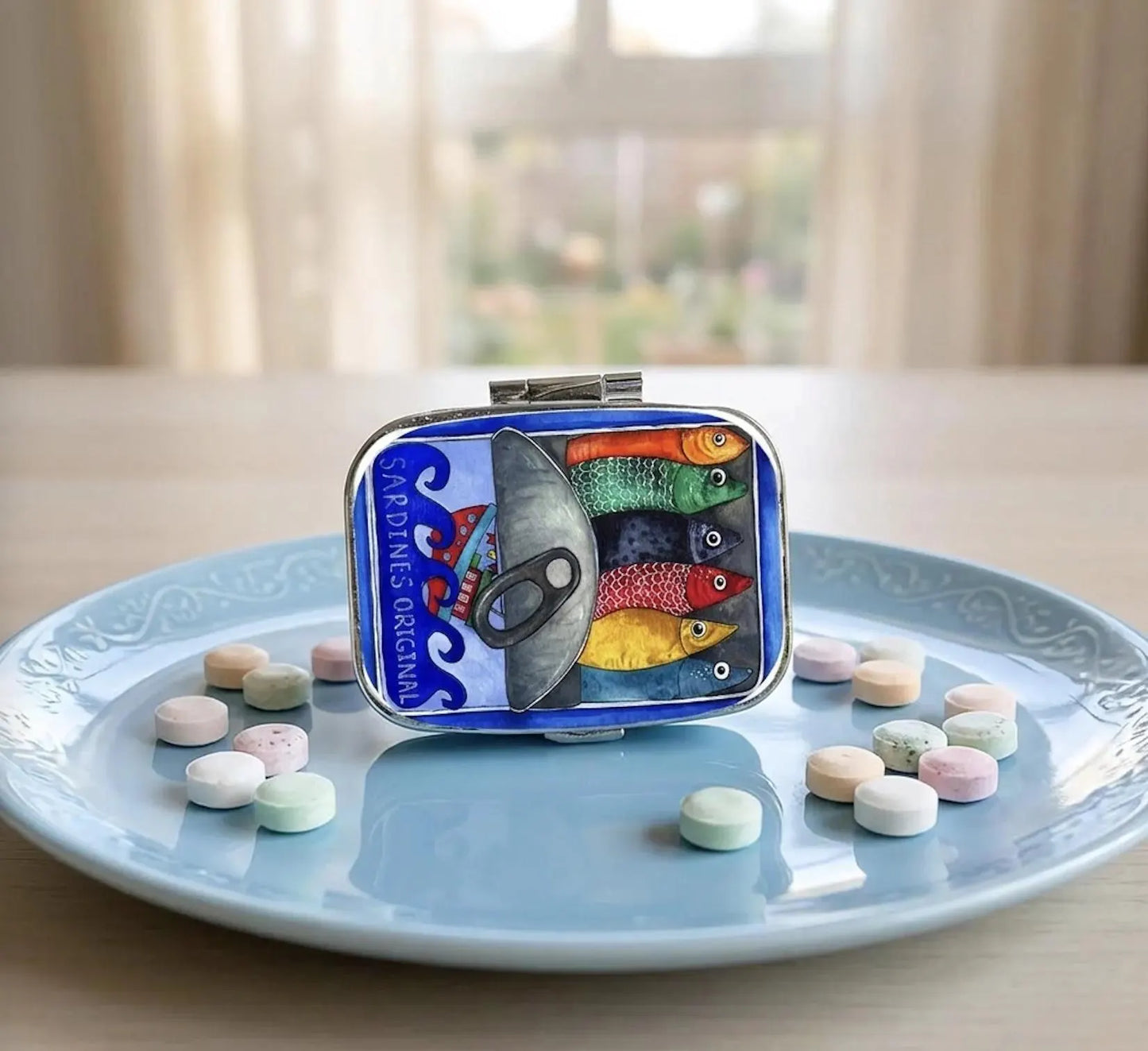 A small rectangular tin with a colorful fish illustration and the text 'PERSIMMA' sits on a light blue plate with scattered pastel candies. The background shows a softly blurred indoor setting with curtains and a table surface. Sardines Pill Case, Pocket Size Metal Mint Holder, Refillable Pill Box, Cute Kawaii Portuguese Art Accessory, Small Gifts, Pill Organizer