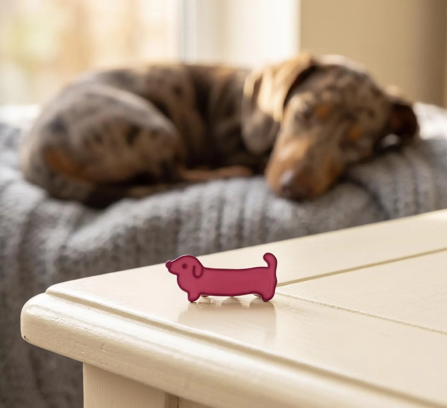 A small pink dachshund-shaped accessory sits on a cream-colored table in the foreground, with a blurred dog resting on a gray blanket in the background. A small pink dachshund-shaped enamel pin rests on a rustic wooden table next to a blue book with gold lettering. The background is a softly blurred living room scene with a plaid armchair and a window. Pink Dachshund Enamel Pin, Cute Wiener Dog Lapel Pin, Dachshund Lover Gift, Doxie Dog Mom Accessories, Hot Pink Sausage Dog Flair, Cute Pins