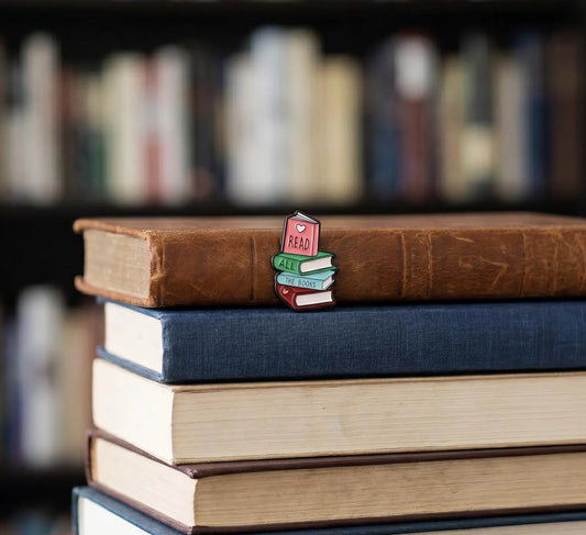 A hand holds a small enamel pin shaped like a stack of books, with the top book reading "READ" and the others reading "MAKE", "LEARN", and "STUDY" in colorful lettering, against a blurred library bookshelf background. Book lover Enamel Pin, Bookworm Lapel Badge, Cute Bookish Gifts, Reader Book Club Accessory, Read all the Books, Bibliophile Gifts, Books