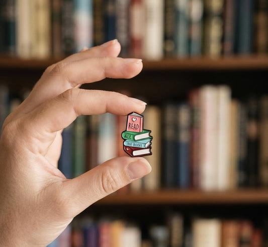 A hand holds a small enamel pin shaped like a stack of books, with the top book reading "READ" and the others reading "MAKE", "LEARN", and "STUDY" in colorful lettering, against a blurred library bookshelf background. Book lover Enamel Pin, Bookworm Lapel Badge, Cute Bookish Gifts, Reader Book Club Accessory, Read all the Books, Bibliophile Gifts, Books