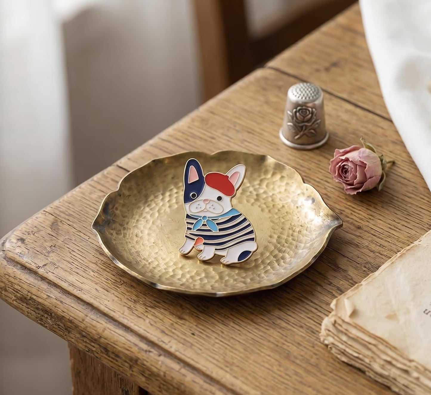 A small enamel lapel pin shaped like a French bulldog wearing a red beret and navy-and-white striped sweater, displayed on a hammered gold dish on a rustic wooden table with a thimble and dried rose nearby. French Bulldog Enamel Pin, Cute Frenchie in Beret Lapel Pin, Dog lovers, Aesthetic Animal Brooch, Parisian Style, Gift for Dog Mom, Cute Pin French Bulldog Enamel Pin, Cute Frenchie in Beret Lapel Pin, Parisian Dog Lover Gift, Dog Mom Backpack Flair, Aesthetic Animal Brooch Gift