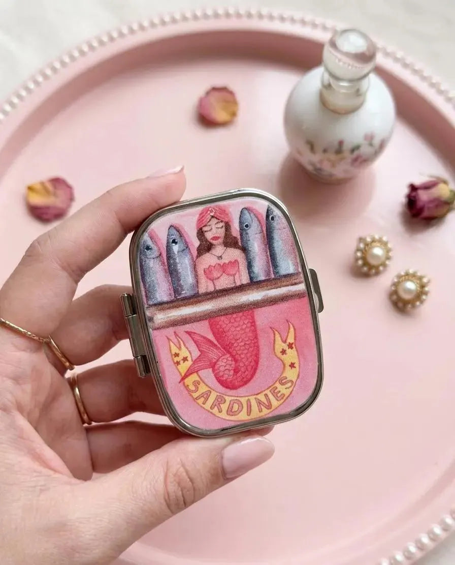A hand holds a small rectangular compact or case with a colorful mermaid and fish illustration and the word 'SARDINES' on the front, set against a pink tray with decorative props. Mermaid Sardines Pill Case, Pocket Size Metal Mint Holder, Refillable Pill Box, Cute Kawaii Accessory, Small Gift Idea