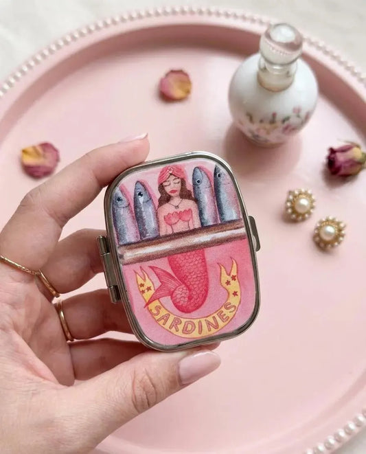 A hand holds a small rectangular compact or case with a colorful mermaid and fish illustration and the word 'SARDINES' on the front, set against a pink tray with decorative props. Mermaid Sardines Pill Case, Pocket Size Metal Mint Holder, Refillable Pill Box, Cute Kawaii Accessory, Small Gift Idea