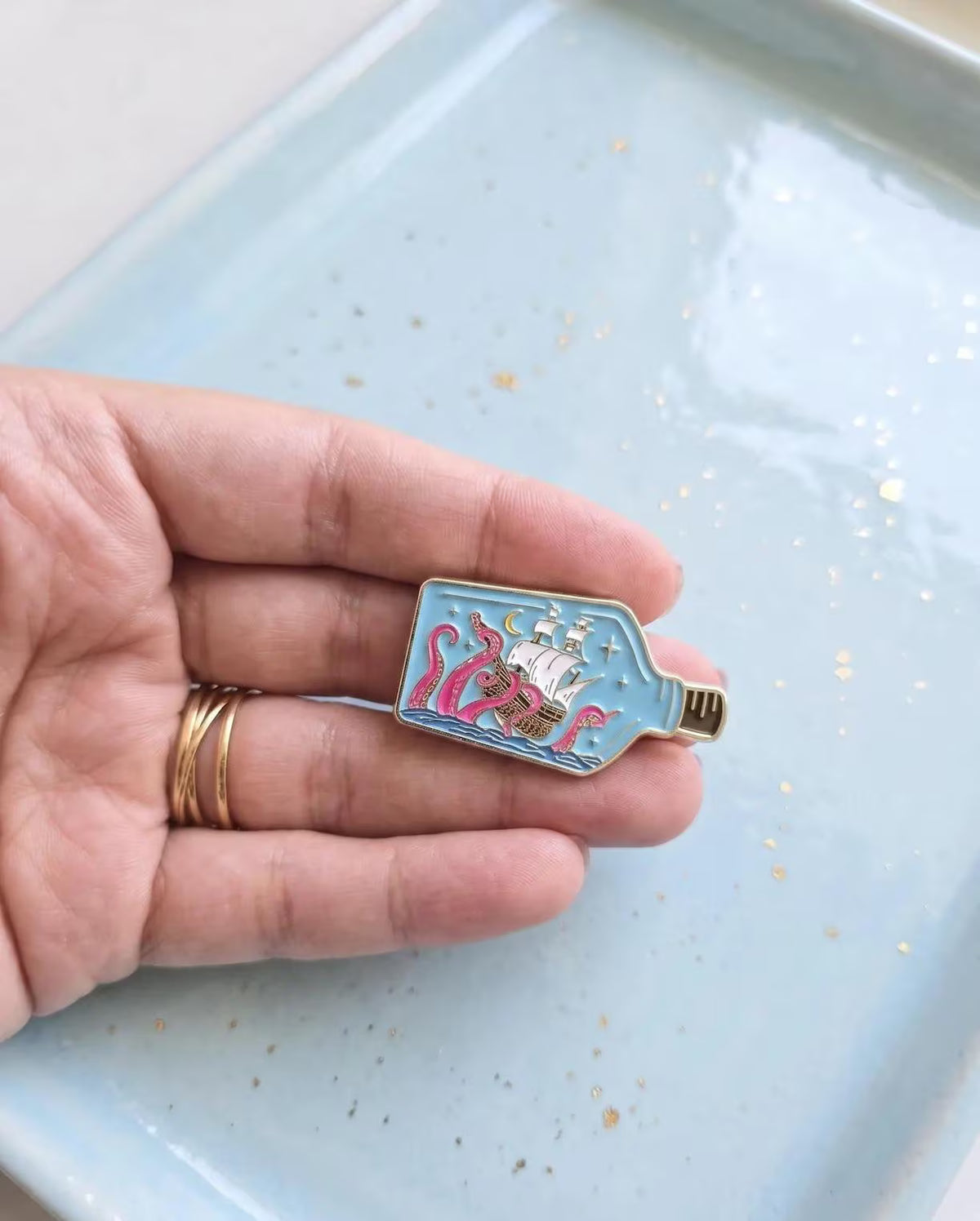 A small enamel pin shaped like a bottle containing a sailing ship and pink sea monsters, resting on a light blue ceramic plate. Kraken Ship in a Bottle Enamel Pin - Nautical Sea Monster Pin - Ocean Kraken Gift - Collectable Pin, Fantasy Lovers - Ocean Lover Gifts