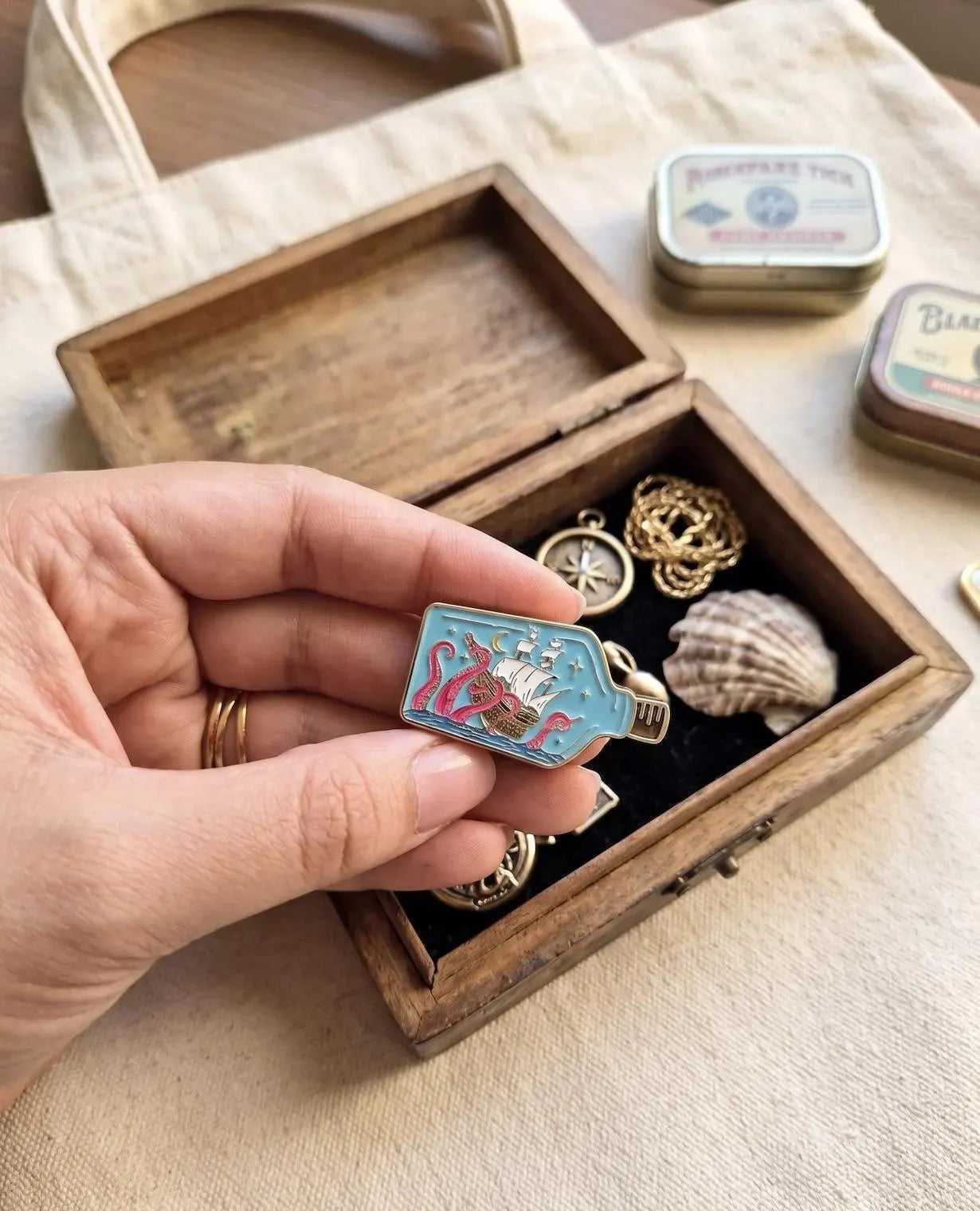 A small enamel pin shaped like a bottle containing a sailing ship and pink sea monsters, resting on a light blue ceramic plate. Kraken Ship in a Bottle Enamel Pin - Nautical Sea Monster Pin - Ocean Kraken Gift - Collectable Pin, Fantasy Lovers - Ocean Lover Gifts