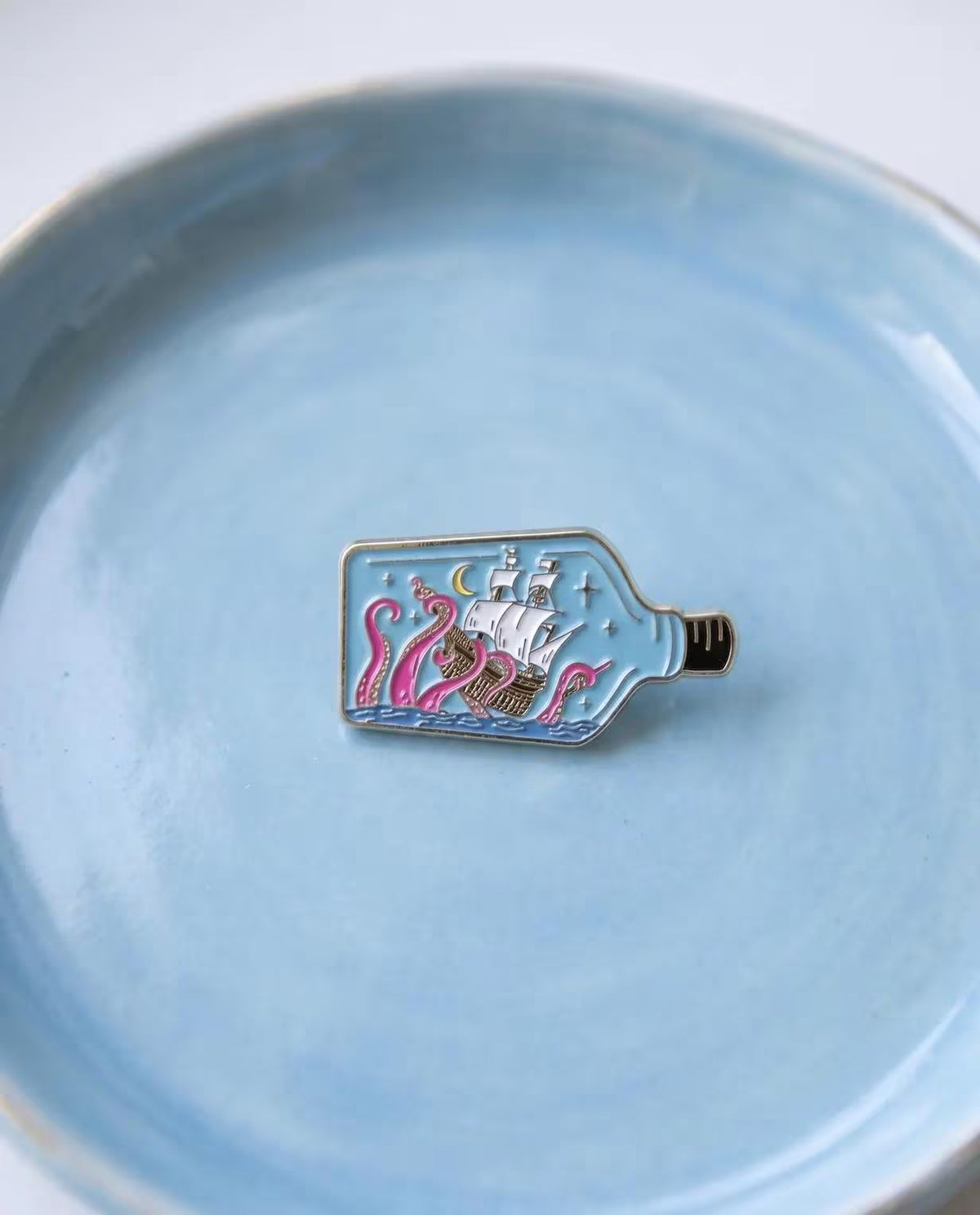 A small enamel pin shaped like a bottle containing a sailing ship and pink sea monsters, resting on a light blue ceramic plate. Kraken Ship in a Bottle Enamel Pin - Nautical Sea Monster Pin - Ocean Kraken Gift - Collectable Pin, Fantasy Lovers - Ocean Lover Gifts