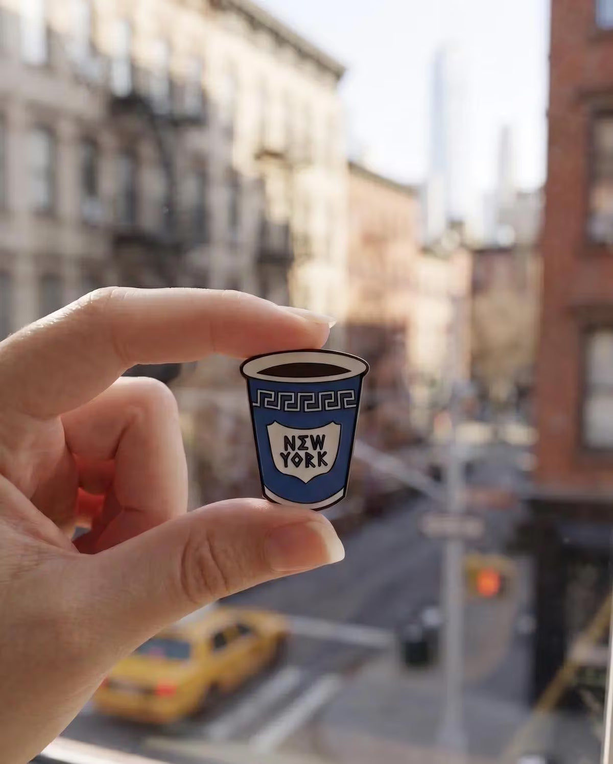 A hand holds a small enamel pin shaped like a blue coffee cup with the words 'NEW YORK' on it, set against a warm, softly blurred cafe table scene with a cup of coffee, spoon, and books. Iconic NYC Blue Coffee Cup Enamel Pin - Anthora Greek Cup Pin, New York City Souvenir, Retro Bodega Aesthetic, Coffee Lover Gifts, Java NYC Coffee Cup Enamel Pin, Iconic Anthora Greek Cup Badge, New York Souvenir Gift, Retro Bodega Aesthetic Pin, Coffee Lover Backpack Flair