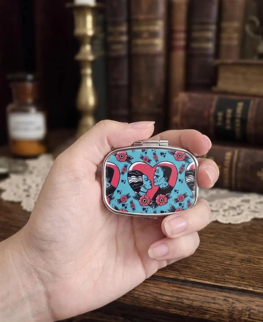 A hand holding a small rectangular metal case with a colorful pattern featuring a pink heart, black and white zebra stripes, and red flowers on a light blue background, set against a blurred background of vintage books and a wooden table. Bride of Frankenstein Pill Box Organizer, Goth Travel Pill Case for Purse, Spooky Horror Gift for Her, Aesthetic Gothic Jewelry Pill Box  Bride of Frankenstein Pill Case, Goth Travel Pill Box for Purse, Spooky Horror Movie Gift, Aesthetic Gothic Mint Tin, Halloween Gift fo