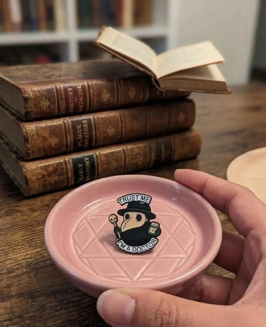 A hand holds a small pink ceramic dish with a printed plague doctor illustration and the words 'TRUST ME ALL DOGS DO TRUST ME' in front of a stack of old books on a wooden table. Plague Doctor Enamel Pin, Goth Kawaii Pin, Trust Me I’m A Doctor, Spooky Collectible, Cute Gift for Friend