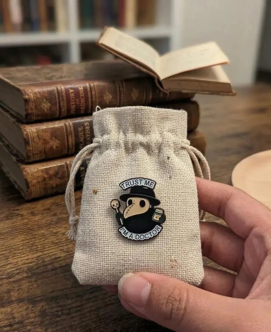 A hand holds a small pink ceramic dish with a printed plague doctor illustration and the words 'TRUST ME ALL DOGS DO TRUST ME' in front of a stack of old books on a wooden table. Plague Doctor Enamel Pin, Goth Kawaii Pin, Trust Me I’m A Doctor, Spooky Collectible, Cute Gift for Friend