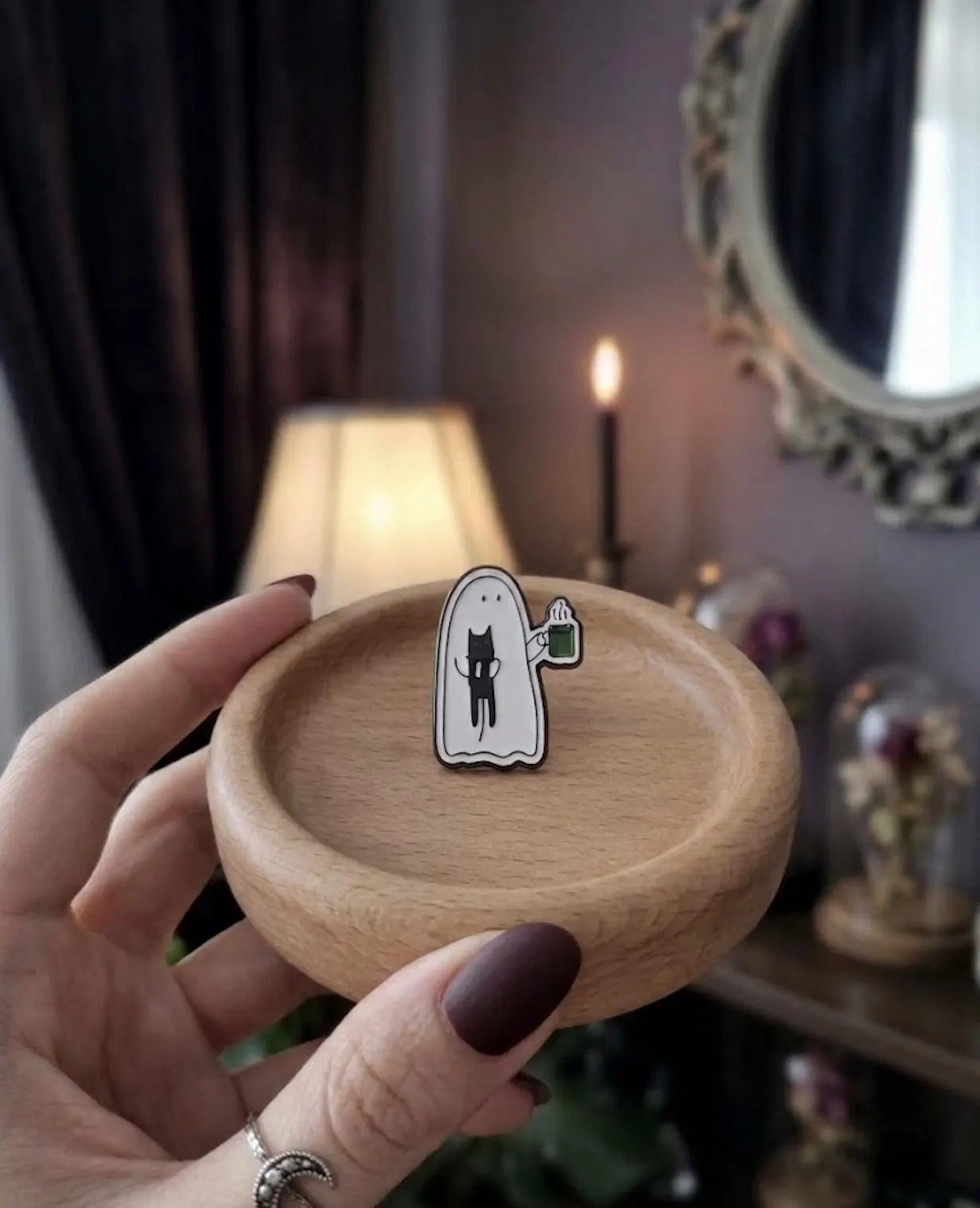 A hand with dark brown manicured nails holds a small enamel pin shaped like a white ghost with a black cat and a green mug, set against a cozy, dimly lit room background with a lamp, candle, and mirror. Cute Ghost Enamel Pin, Soft Goth Black Cat Lapel Pin, Spooky Cute Coffee Break Matcha Mug, Kawaii Halloween Flair Gift for Cat Lovers