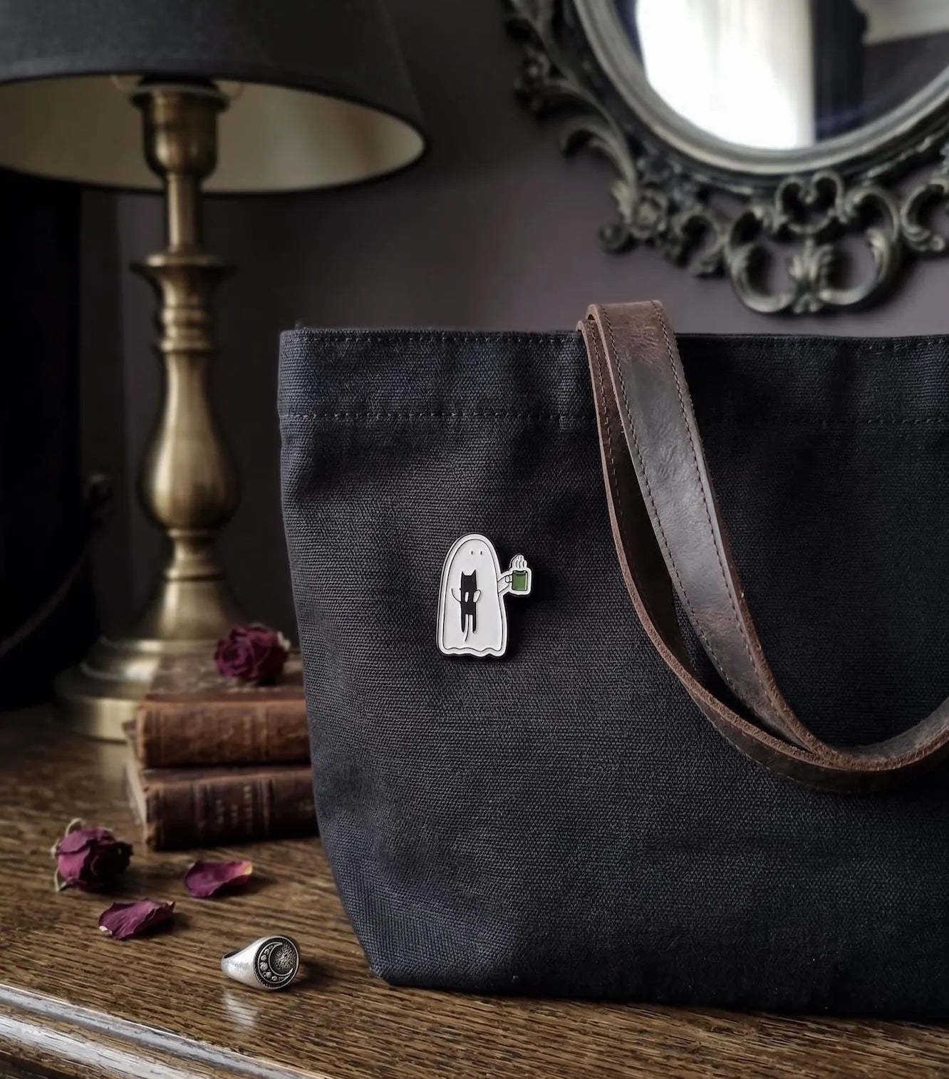 A hand with dark brown manicured nails holds a small enamel pin shaped like a white ghost with a black cat and a green mug, set against a cozy, dimly lit room background with a lamp, candle, and mirror. Cute Ghost Enamel Pin, Soft Goth Black Cat Lapel Pin, Spooky Cute Coffee Break Matcha Mug, Kawaii Halloween Flair Gift for Cat Lovers