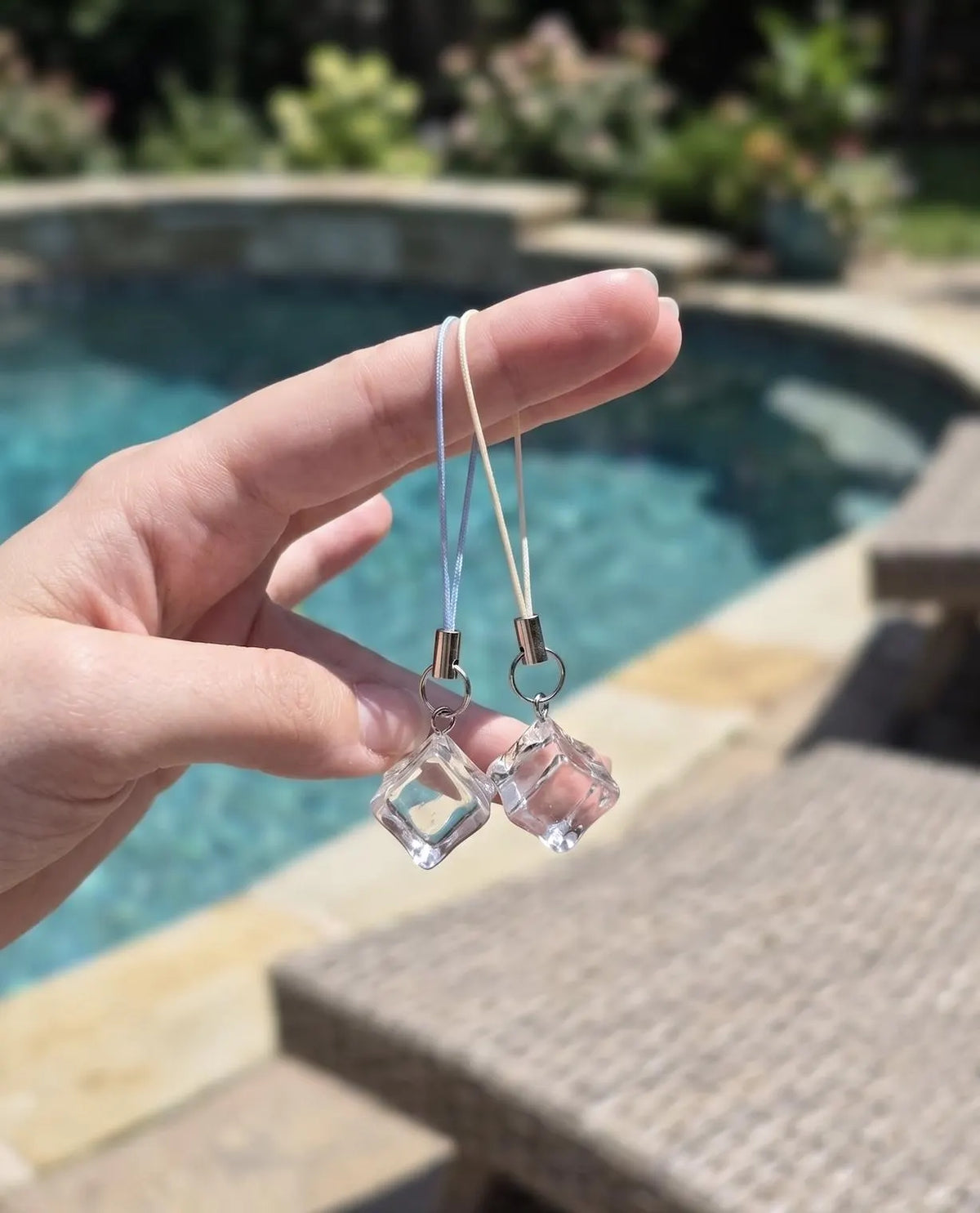 A hand holds a pair of clear faceted cube dangle earrings on thin white cords, photographed outdoors with a swimming pool and patio furniture blurred in the background. Minimalist Refreshing Ice Cube Phone Charm, Summer Aesthetic Clear Charm, Cute Phone Accessory, Clear Resin Ice Phone Strap