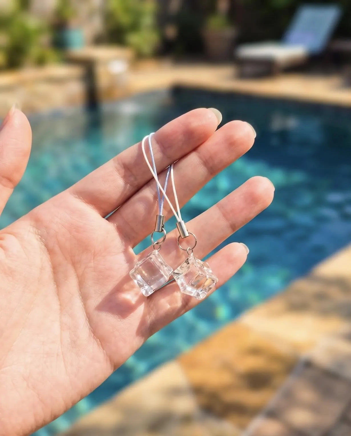 A hand holds a pair of clear faceted cube dangle earrings on thin white cords, photographed outdoors with a swimming pool and patio furniture blurred in the background. Minimalist Refreshing Ice Cube Phone Charm, Summer Aesthetic Clear Charm, Cute Phone Accessory, Clear Resin Ice Phone Strap