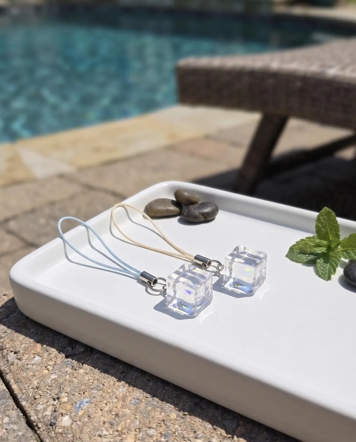 A hand holds a pair of clear faceted cube dangle earrings on thin white cords, photographed outdoors with a swimming pool and patio furniture blurred in the background. Minimalist Refreshing Ice Cube Phone Charm, Summer Aesthetic Clear Charm, Cute Phone Accessory, Clear Resin Ice Phone Strap