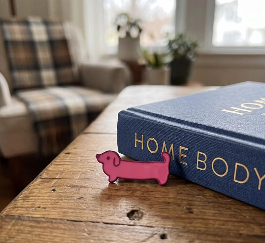 A small pink dachshund-shaped enamel pin rests on a rustic wooden table next to a blue book with gold lettering. The background is a softly blurred living room scene with a plaid armchair and a window. Pink Dachshund Enamel Pin, Cute Wiener Dog Lapel Pin, Dachshund Lover Gift, Doxie Dog Mom Accessories, Hot Pink Sausage Dog Flair, Cute Pins