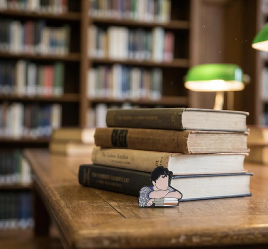 A small enamel pin shaped like a cartoon girl resting her head on stacked books, placed on a wooden table in a blurred library setting with bookshelves and a green lamp in the background. Contemplating Reader Enamel Pin, Book Lover Pin, Reading, Bibliophile, thinking pin, Bookworm Gift, Bookish Writers Gift, Dark Academia