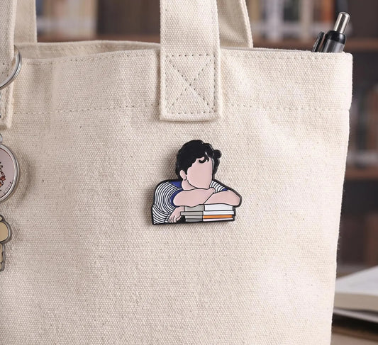 A close-up of a beige canvas tote bag with a cartoon enamel pin of a person resting their head on books. A pen is tucked into the side pocket, and the background is a softly blurred indoor setting. A small enamel pin shaped like a cartoon girl resting her head on stacked books, placed on a wooden table in a blurred library setting with bookshelves and a green lamp in the background. Contemplating Reader Enamel Pin, Book Lover Pin, Reading, Bibliophile, thinking pin, Bookworm Gift, Bookish Writers Gift, Dark