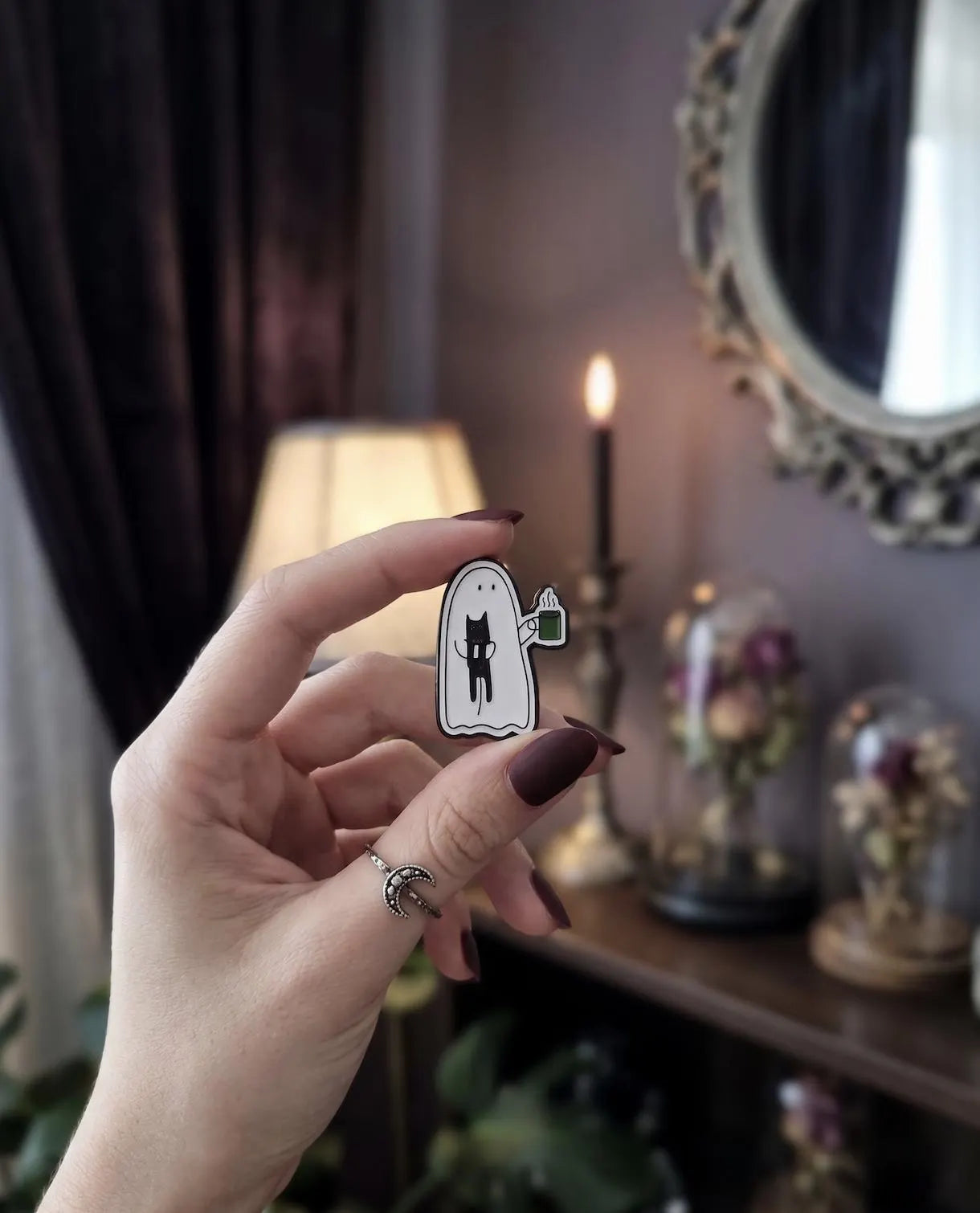 A hand with dark brown manicured nails holds a small enamel pin shaped like a white ghost with a black cat and a green mug, set against a cozy, dimly lit room background with a lamp, candle, and mirror. Cute Ghost Enamel Pin, Soft Goth Black Cat Lapel Pin, Spooky Cute Coffee Break Matcha Mug, Kawaii Halloween Flair Gift for Cat Lovers