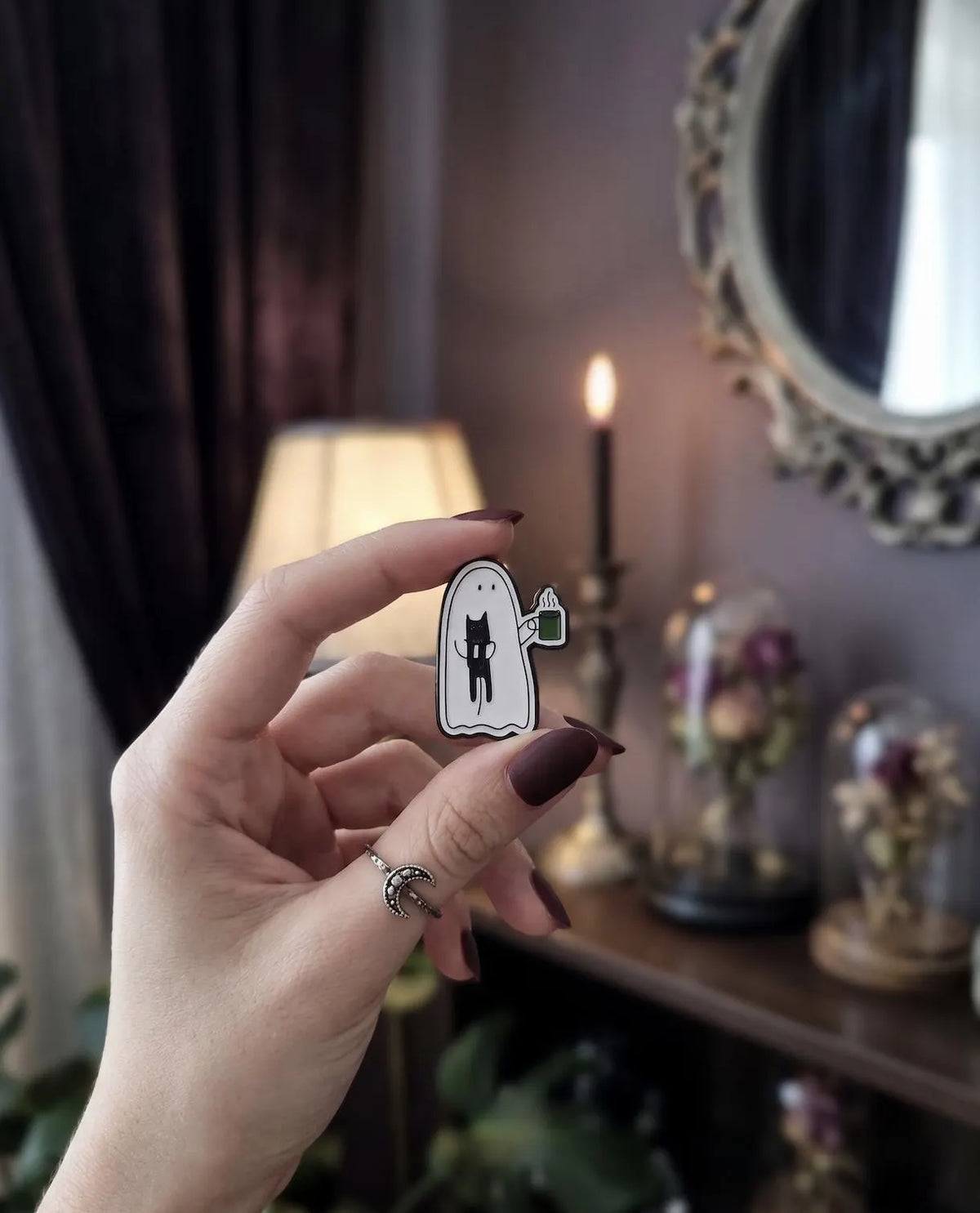 A hand with dark brown manicured nails holds a small enamel pin shaped like a white ghost with a black cat and a green mug, set against a cozy, dimly lit room background with a lamp, candle, and mirror. Cute Ghost Enamel Pin, Soft Goth Black Cat Lapel Pin, Spooky Cute Coffee Break Matcha Mug, Kawaii Halloween Flair Gift for Cat Lovers