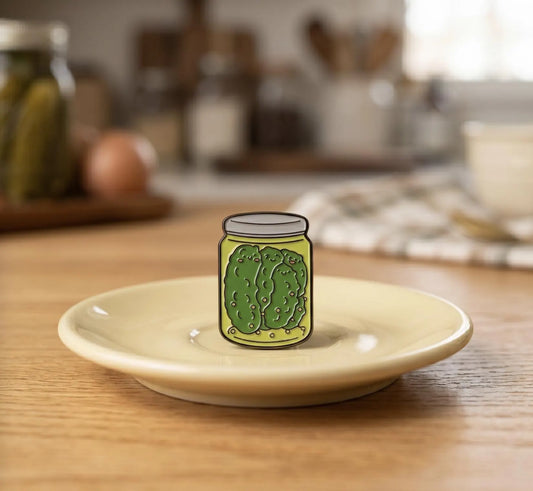 A hand holds a small enamel pin shaped like a pickle jar with a green pickle illustration, set against a blurred background of glass pickle jars on a wooden shelf. Cute Pickle Family Enamel Pin | Kawaii Dill Jar Lapel Badge | Funny Food Pickle Lover Gift | Pickle in a Jar, Pickle Girl, Foodie Gifts