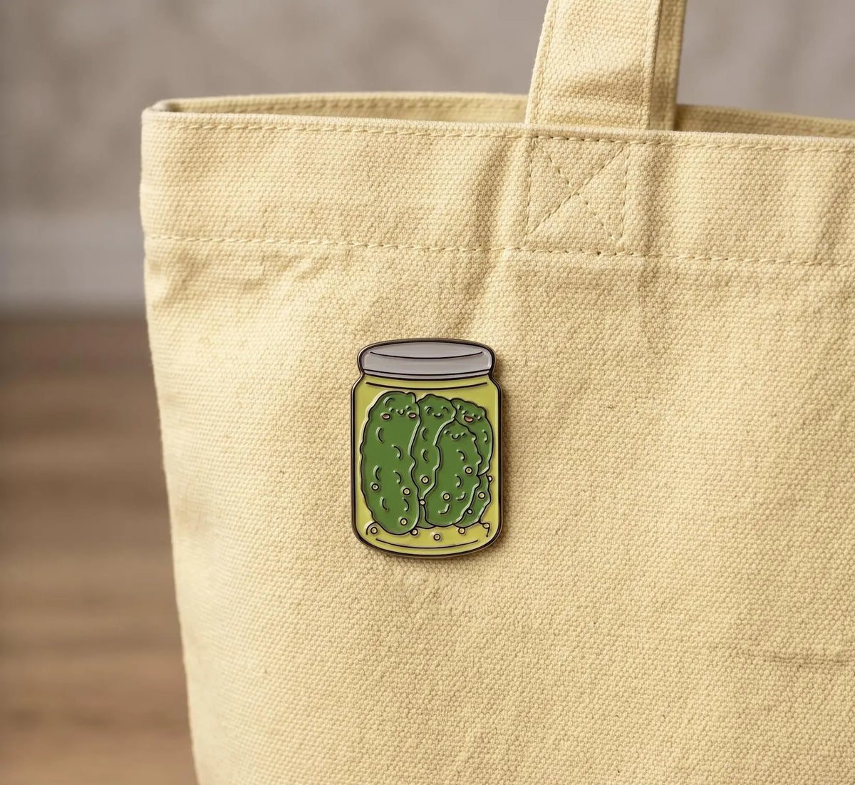 A hand holds a small enamel pin shaped like a pickle jar with a green pickle illustration, set against a blurred background of glass pickle jars on a wooden shelf. Cute Pickle Family Enamel Pin | Kawaii Dill Jar Lapel Badge | Funny Food Pickle Lover Gift | Pickle in a Jar, Pickle Girl, Foodie Gifts