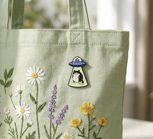 A sage green canvas tote bag with embroidered wildflowers and a small pin featuring a black cat under a UFO, photographed in soft natural light. A small enamel pin shaped like a black-and-white cat under a blue UFO with green rays, displayed on a wooden tray with colorful paper and an ink bottle in the background.Black Cat Alien Enamel Pin - Space Core UFO Cat Pin for Backpacks - Cute Galaxy Kitten Pin - Unique Spooky Gift for Cat Lovers, Cosmic Cat