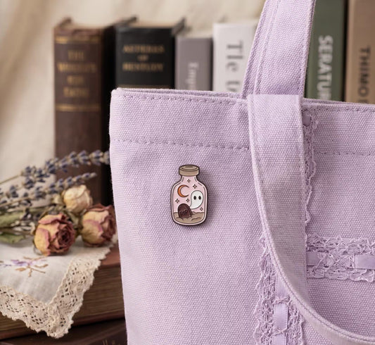 A lavender canvas tote bag with a small bottle-shaped pin featuring a ghost and moon design, styled on a table with dried flowers and books in the background. Cute Ghost in Potion Bottle Enamel Pin, Kawaii Halloween Pin, Soft goth Aesthetic, RIP Tombstone, Gift for spooky book lovers, Pastel Purple