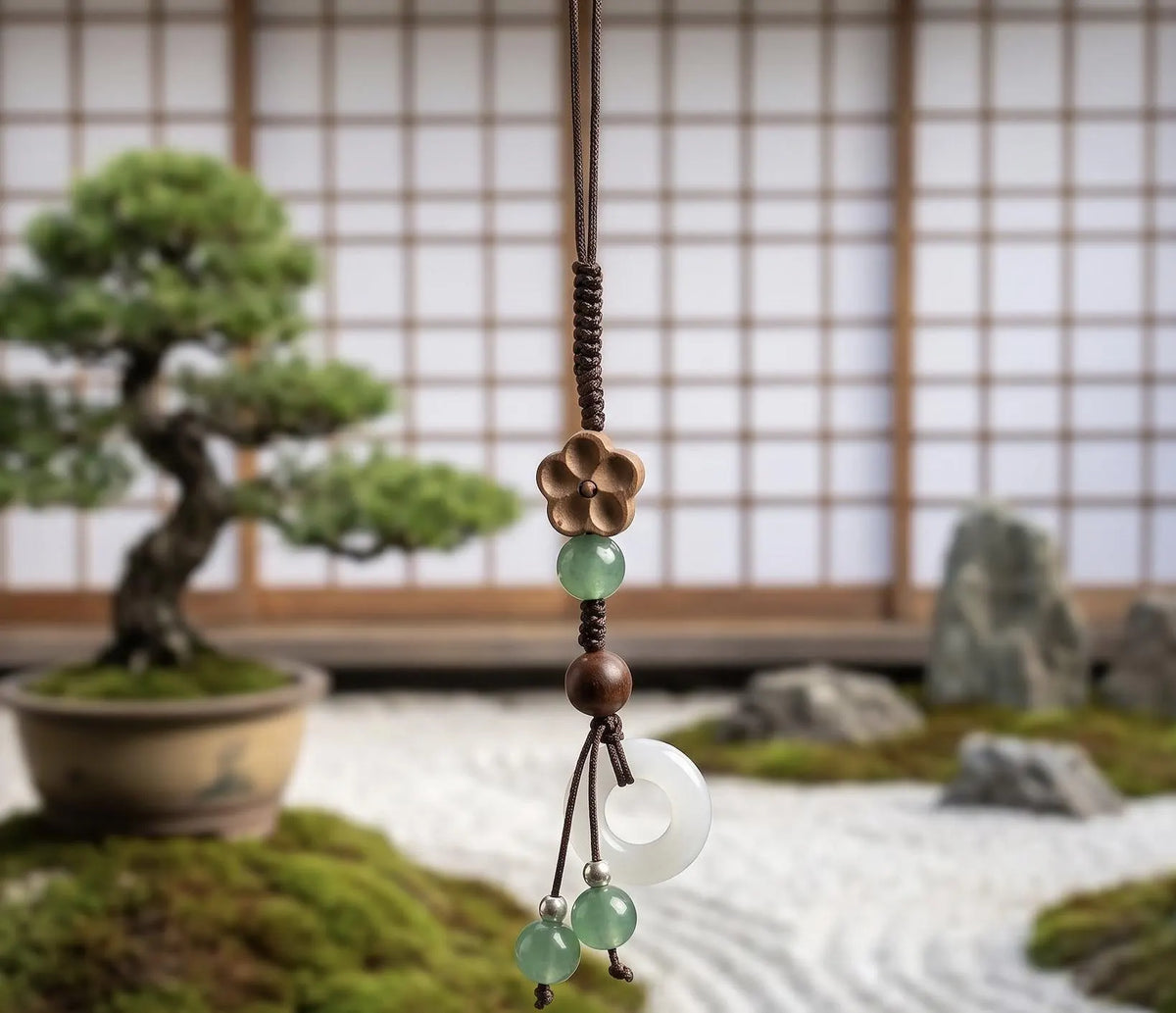 A close-up of a hand holding a black smartphone, showing a decorative phone strap with a braided cord, bronze flower charm, green and brown beads, a white jade disc, and small green beads, set against a softly blurred Japanese-style interior background. Plum Blossom Phone Charm, Wooden Flower Phone Strap, Jade Green Glass Ring Accessory, Cute Aesthetic Bag Flair, Cozy Handmade Gift for Her