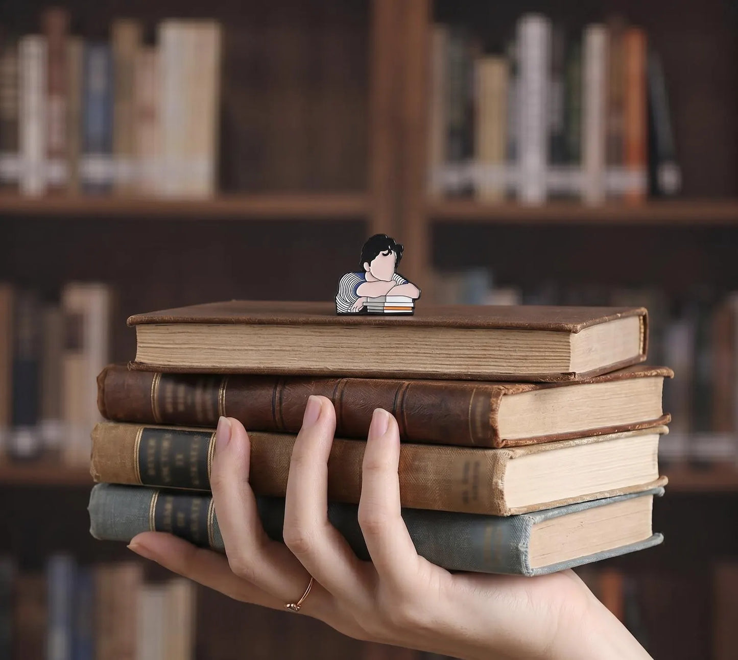 A small enamel pin shaped like a cartoon girl resting her head on stacked books, placed on a wooden table in a blurred library setting with bookshelves and a green lamp in the background. Contemplating Reader Enamel Pin, Book Lover Pin, Reading, Bibliophile, thinking pin, Bookworm Gift, Bookish Writers Gift, Dark Academia