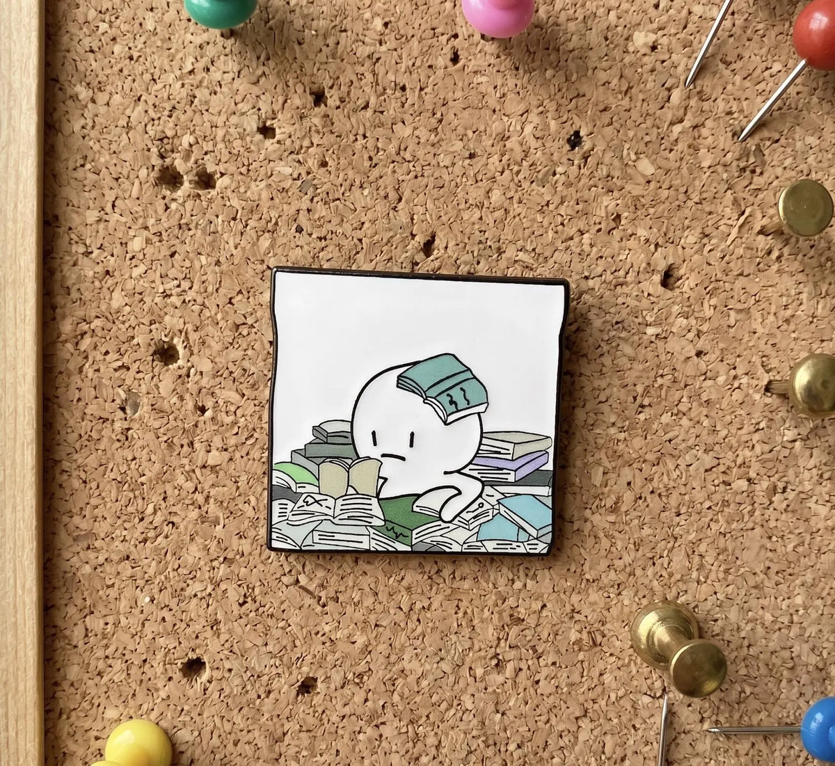 A hand holds a small square charm or pendant featuring a cartoon character surrounded by books, with a softly blurred living room in the background. Too Many Books Too Little Time Enamel Pin, Funny Reading Lapel Pin for Book Lovers, Cute Bookworm Gift, Relatable Bibliophile Reader Bag Pin