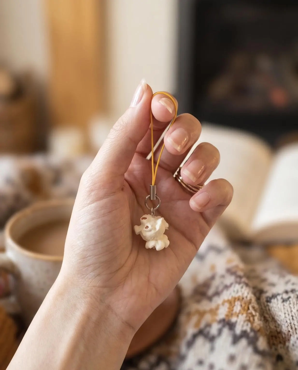 A hand holding a small cream-colored plush charm on a yellow cord, with a cozy indoor background featuring a lamp, books, and a knitted blanket. Popcorn Lover Phone Charm, Cute Miniature Food Keychain, Movie Lover Gift, Funky Cute Keychain, Corn gift, Quirky Movie Theater, Movie Night