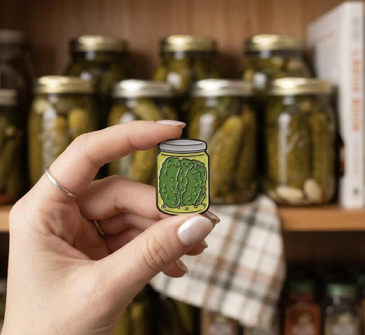 A hand holds a small enamel pin shaped like a pickle jar with a green pickle illustration, set against a blurred background of glass pickle jars on a wooden shelf. Cute Pickle Family Enamel Pin | Kawaii Dill Jar Lapel Badge | Funny Food Pickle Lover Gift | Pickle in a Jar, Pickle Girl, Foodie Gifts